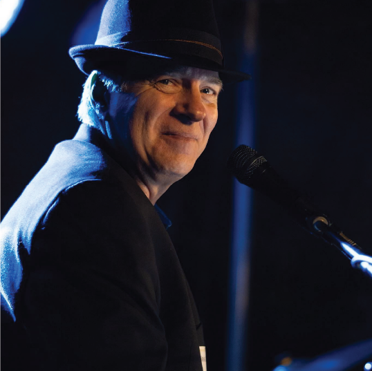 Taras Nahirniak, choral musical composer, smiles while seated at a piano under stage lighting, wearing a black suit and fedora, with a microphone.